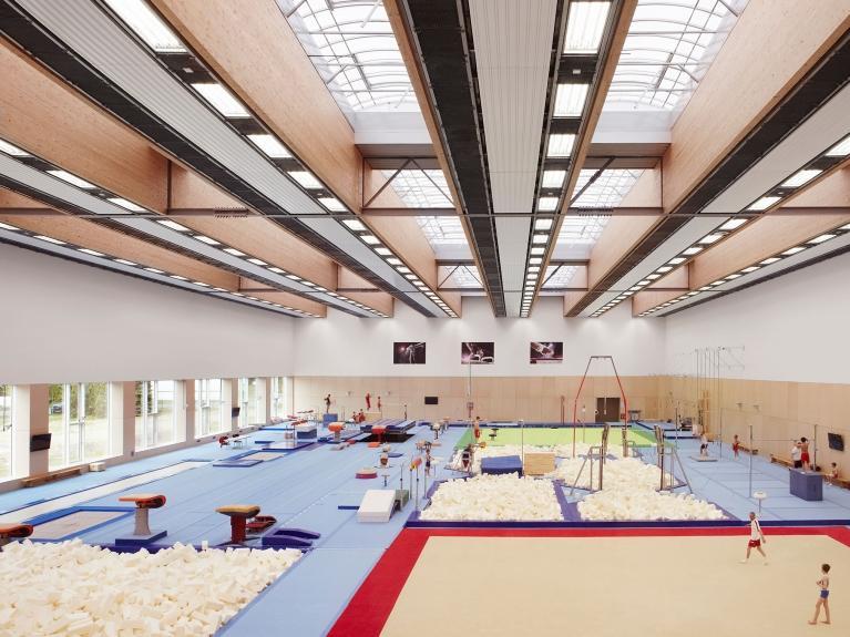 Innenaufnahme einer modernen Turnhalle des olympischen und paralympischen Trainingszentrums in Grünheide mit verschiedenen Turngeräten wie Sprungtische, Schwebebalken und Barren. Realisierung eines klimaneutralen Energiekonzeptes für das Trainingszentrum.
