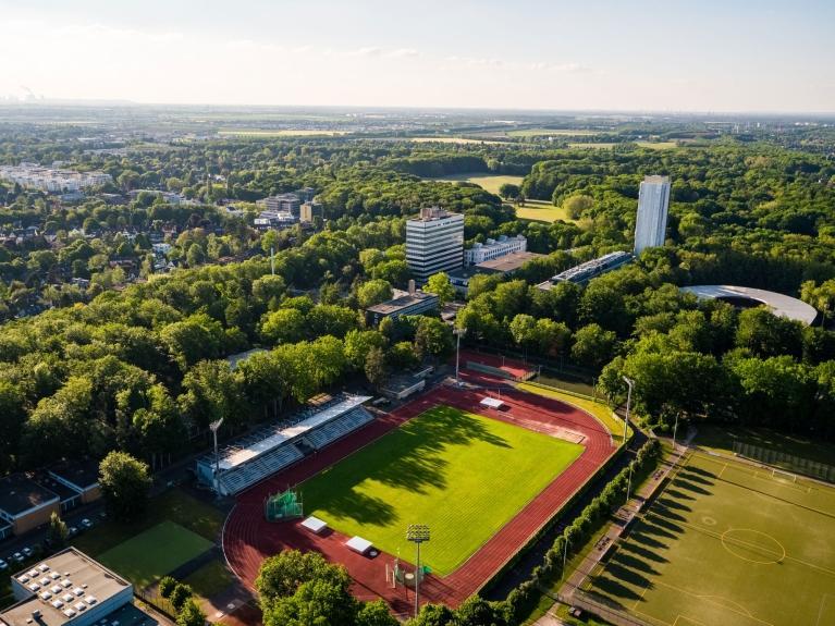 Außenaufnahme des Campus der Deutschen Sporthochschule Köln mit Stadion, Sportanlagen und Gebäuden, umgeben von Bäumen
