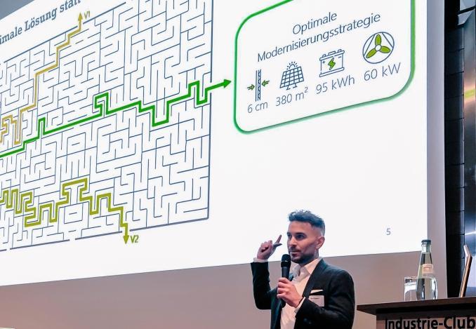 Redner steht vor Pult und vor Screen mit Daten, Zahlen