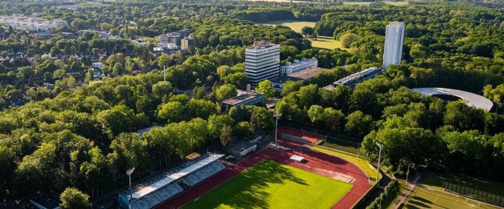 Außenaufnahme des Campus der Deutschen Sporthochschule Köln mit Stadion, Sportanlagen und Gebäuden, umgeben von Bäumen
